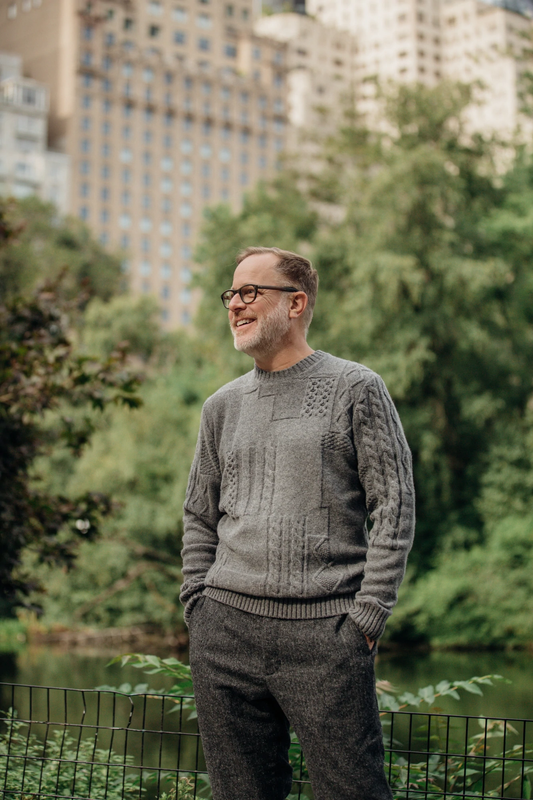 A man stands in a park wearing a grey knit sweater. 