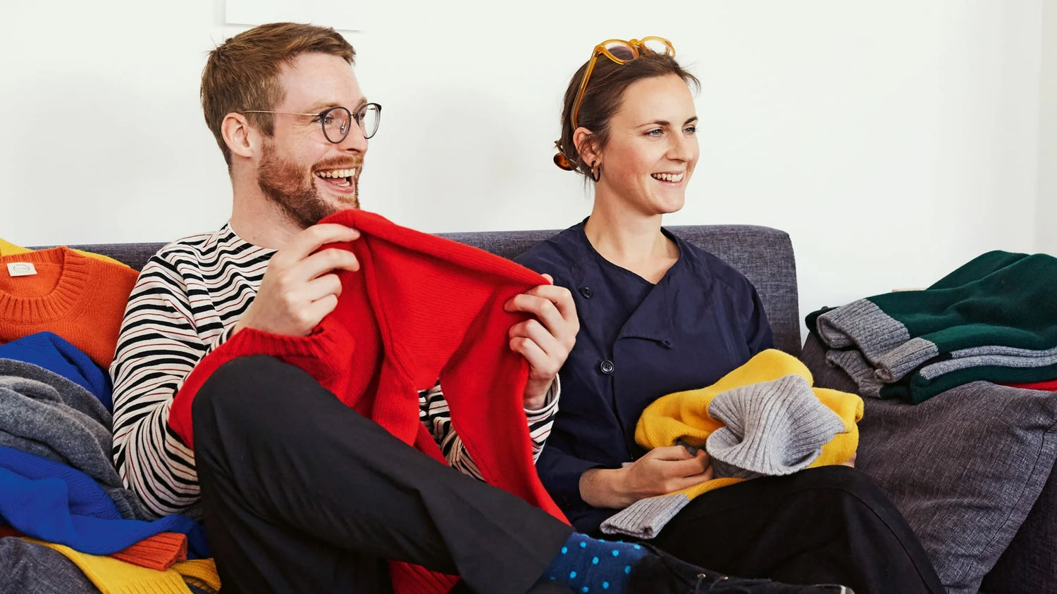 Alice and Ben sit on a couch, holding up colorful knitwear. 