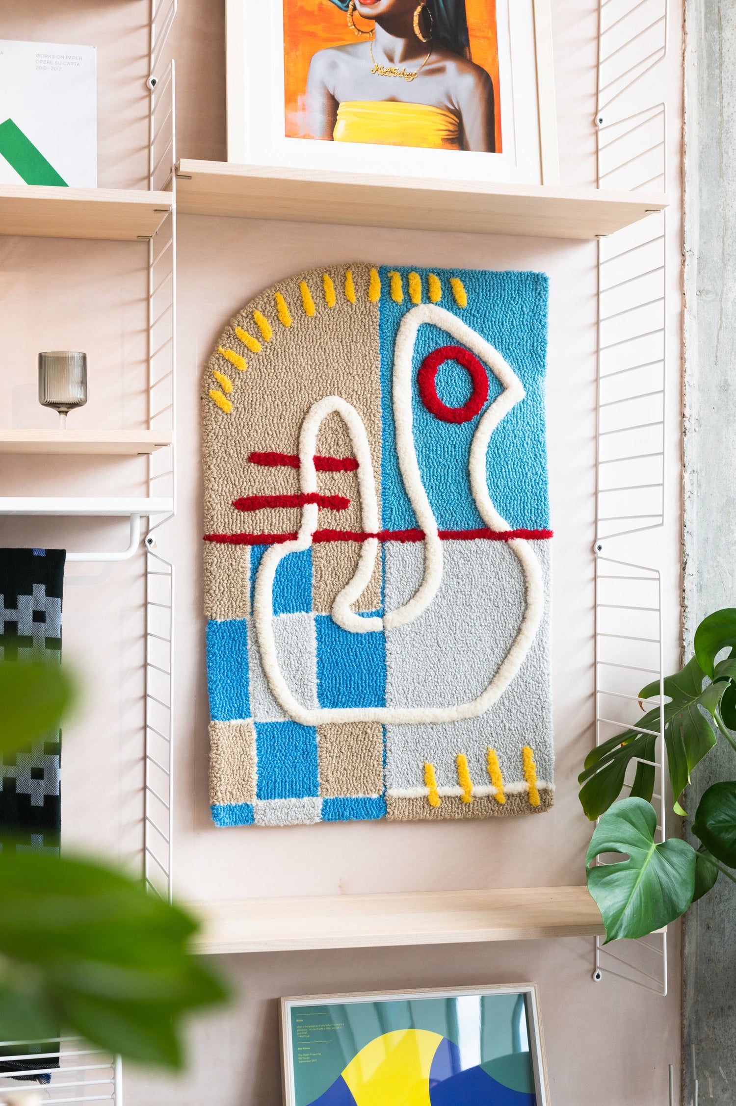 An artistic wool wall hanging shown among plants and shelves.