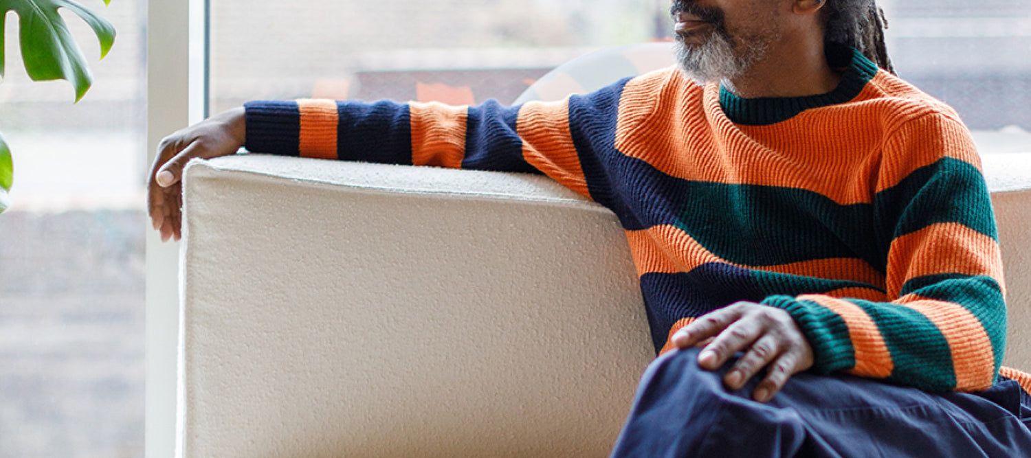 A man wearing a bright orange knit sweater sits on a couch.